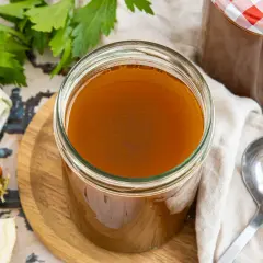 Ein Glas goldbrauner Rinderfond, bereit zum Verfeinern von Gerichten. Daneben Karotte, Lauch und Petersilie – die Zutaten, um Rinderfond selber machen zu können.