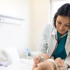 Lekarka w białym fartuchu osłuchuje stetoskopem noworodka leżącego na łóżku.