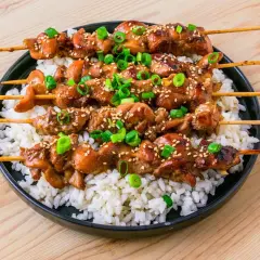 Fyra kycklingspett teriyaki ligger på en bädd av vitt ris, toppade med sesamfrön och hackad salladslök.