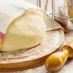 Masa de pan cubierta con un paño a rayas, sobre una tabla de madera con harina esparcida. Rodillo y tamizador de harina, ingredientes para hacer pan.
