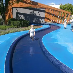 Chłopiec pluska się w wodzie na niebieskiej nawierzchni w parku bajka w Błoniu. Obok idzie kobieta z dzieckiem.