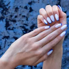 Eleganckie, białe paznokcie z granatowym french manicure, prezentowane na tle niebieskiego, aksamitnego materiału.