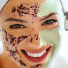 Una mujer sonríe con una mascarilla verde y un exfoliante de café y azul en su rostro.