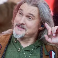 Maciej, wokalista grupy Dżem, z długimi siwymi włosami i brodą, w zielonej koszuli i kamizelce, poprawia słuchawkę w uchu.