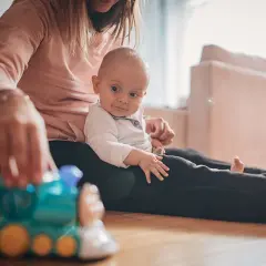 Mama bawi się z 9-miesięcznym dzieckiem, które siedzi na jej kolanach. Przed nimi leży zabawkowy pociąg, idealny do zabawy dla malucha.