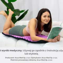 Kobieta czyta książkę, leżąc na fioletowej macie do akupresury na brzuch. Relaks i zdrowie.