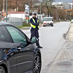 Policjant zatrzymuje czarne auto, być może za recydywę mandatu. Na drodze stoi biały van.