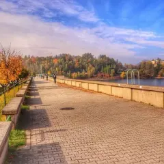 Jesioro Czartoryskie jesienią. Spacerowicze i rowerzyści na promenadzie, w tle zamek na wyspie i kolorowe drzewa.