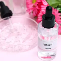 Serum z kwasem mlekowym w szklanej buteleczce z pipetą, obok różowych kwiatów.