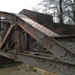 Zardzewiały, nitowany most w awinionie, z widocznymi elementami konstrukcyjnymi i budynkiem w tle.