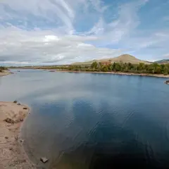 El embalse del Pontón, con aguas azules y orillas arenosas, se extiende hacia colinas verdes y un edificio de ladrillo rojo bajo un cielo nublado.