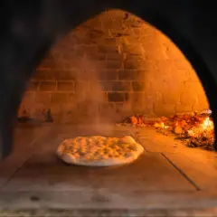 Wnętrze pieca do pizzy z palącym się drewnem i surowym ciastem na kamiennej podłodze. Dym unosi się wokół.
