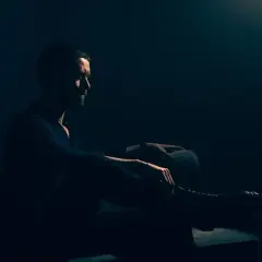 Mężczyzna z brodą siedzi w półmroku, wpatrzony w dal. Jego postawa jest **low key**, emanuje spokojem i zadumą.