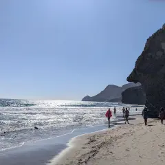 Gente pasea por la **playa de Mónsul** bajo un cielo azul, con olas rompiendo en la orilla y una gran roca a la derecha.