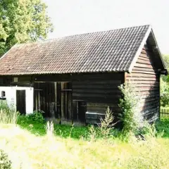 Drewniany budynek gospodarczy z ciemną elewacją i dachówką, otoczony zielenią.