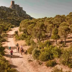 Randonneurs et cavaliers sur le sentier cathare GR 367, menant vers un château perché sur la montagne. Paysage aride et boisé.