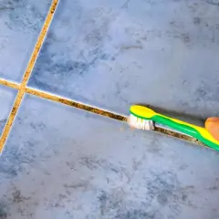 Main tenant, une brosse à dents verte et jaune est utilisée pour nettoyer joint salle de bain, révélant des carreaux bleus.