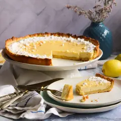 Eine köstliche Tarte au Citron, bestäubt mit Puderzucker, steht bereit. Ein Stück wurde bereits auf einem Teller serviert, daneben liegen frische Zitronen.
