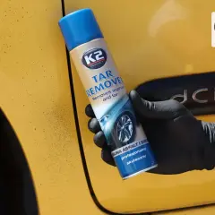 Rękawiczka z czarną gumą trzyma puszkę preparatu K2 Tar Remover, który skutecznie usuwa smołę z żółtego lakieru samochodu.