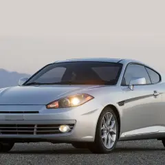 Srebrny Hyundai Tuscani na tle górskiego krajobrazu. Sportowy samochód z błyszczącymi felgami i włączonymi światłami.