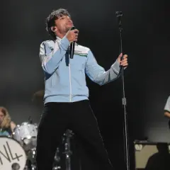 Louis Tomlinson na scenie, śpiewający do mikrofonu. W tle perkusista i gitarzysta.