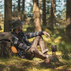 Man med hatt och rutig skjorta vilar vid ett träd under en vandring i Småland. Ryggsäck och stormkök redo för äventyr.