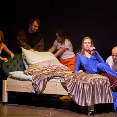Pięć osób na scenie, w tym kobieta w niebieskiej sukience z mikrofonem, przy łóżku.