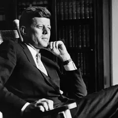 John F. Kennedy sitzt nachdenklich in einem Schaukelstuhl, seine Hand am Kinn. Trotz seiner bekannten **John F. Kennedy Krankheit** strahlt er eine ruhige Entschlossenheit aus.