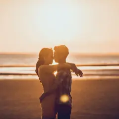 Para zakochanych na plaży o zachodzie słońca. Mężczyzna patrzy kobiecie na usta, obejmując ją.