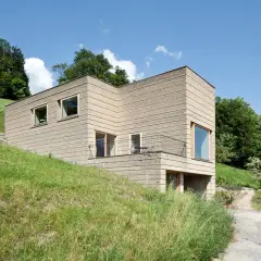 Modernes Haus mit rauer Fassade, das die natürliche Textur von **Lehm als Baustoff** zeigt. Eingebettet in eine grüne Hügellandschaft unter blauem Himmel.
