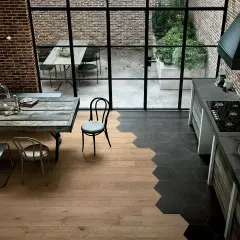 Cucina con stacco tra parquet e piastrelle esagonali nere. Tavolo rustico, sedie, mattoni a vista e grande finestra sul cortile.