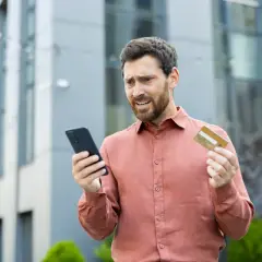 Mężczyzna z kartą płatniczą i telefonem, wyrażający zaniepokojenie.