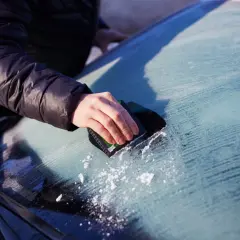 En hand skrapar is från insidan av bilrutan. Snöflingor syns på den frostiga ytan.