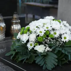 Nowoczesne kompozycje ze sztucznych kwiatów na nagrobku, obok zniczy. Bukiet z białych chryzantem i zielonych kulek.