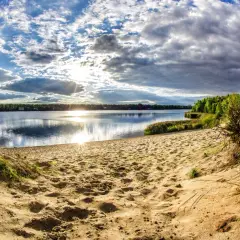 Piaskowa plaża nad jeziorem, słońce przebija się przez chmury, tworząc refleksy na wodzie. W oddali las, a po prawej stronie widoczne korzenie drzew. Idealne miejsce na wypoczynek nad pogorią zalew.