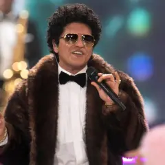 Bruno Mars w futrzanym płaszczu i okularach przeciwsłonecznych śpiewa na koncercie, trzymając mikrofon.