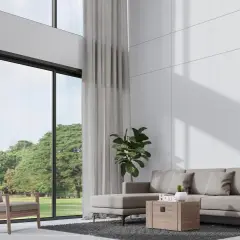 Nowoczesny salon z wysokim sufitem, szara sofa, fotel i wielkie okna z widokiem na zieleń.