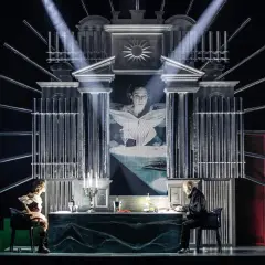 Scena teatralna z projekcją twarzy kobiety, dwójką siedzących przy stole postaci i stojącym mężczyzną. Atmosfera pełna jest teatralnych misteriów.