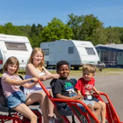 Fyra glada barn på en trampbil på Kolmården first camp. Husvagnar och träd i bakgrunden.