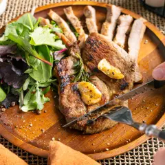 Ein saftiges Schweinesteak in der Pfanne, mit Rosmarin und Knoblauch verfeinert, wird mit einem Messer und einer Gabel angeschnitten. Dazu gibt es einen frischen Salat.