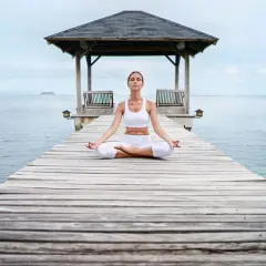 Frau in weißer Kleidung meditiert auf einem Steg am Meer. Eine Anleitung zur transzendentalen Meditation, um inneren Frieden zu finden.