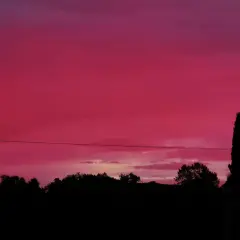 Un ciel rouge le soir, teinté de rose, surplombe une silhouette d'arbres et d'une maison.