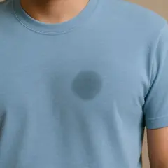 Ein Mann trägt ein blaues T-Shirt mit einem dunklen Fleck auf der Brust. Tipps, wie man fettflecken aus kleidung entfernen nach dem waschen kann.