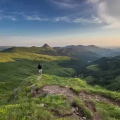 Un randonneur contemple un paysage de montagnes verdoyantes, un panorama à couper le souffle. Le soleil baigne les collines d'une lumière dorée, créant des ombres profondes. Le sentier sinueux, d'une longueur d'environ 400 mètres, invite à l'exploration.