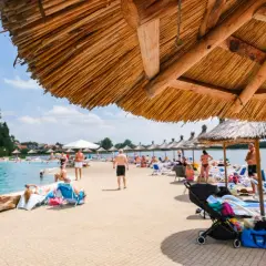 Słoneczny dzień nad wodą, ludzie odpoczywają pod strzechami. Sprawdź molo Osiek cennik i zaplanuj swój relaks!