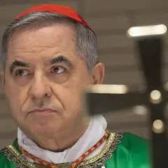 Kardinal Becciu trägt eine rote Pileolus und eine grüne Soutane. Ein Kreuz ist im Vordergrund unscharf.