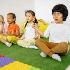 Kinder üben Achtsamkeit im Unterricht, sitzen im Schneidersitz und meditieren. Ein Junge im Vordergrund führt die Übung an.