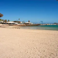 Para relaksuje się na plaży playa del castillo, ciesząc się słońcem i lodami. W tle widać turkusowe morze i zabudowania portowe.