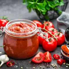 Tarro de cristal lleno de vibrante salsa de tomate casera, rodeado de tomates frescos, ajos, guindillas y especias.