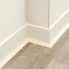 Angolo di un battiscopa in ceramica bianca, pronto per essere incollato su un pavimento in laminato grigio.